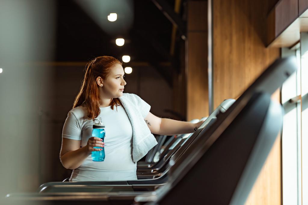 selective focus of overweight girl with towel on shoulder holding sports bottle while standing at treadmill - Photo, Image