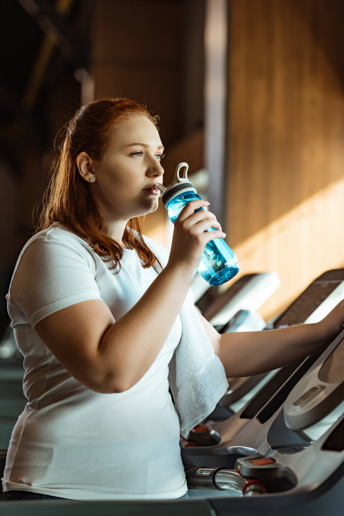 pensive overweight girl drinking from sports bottle while standing at treadmill - Photo, Image