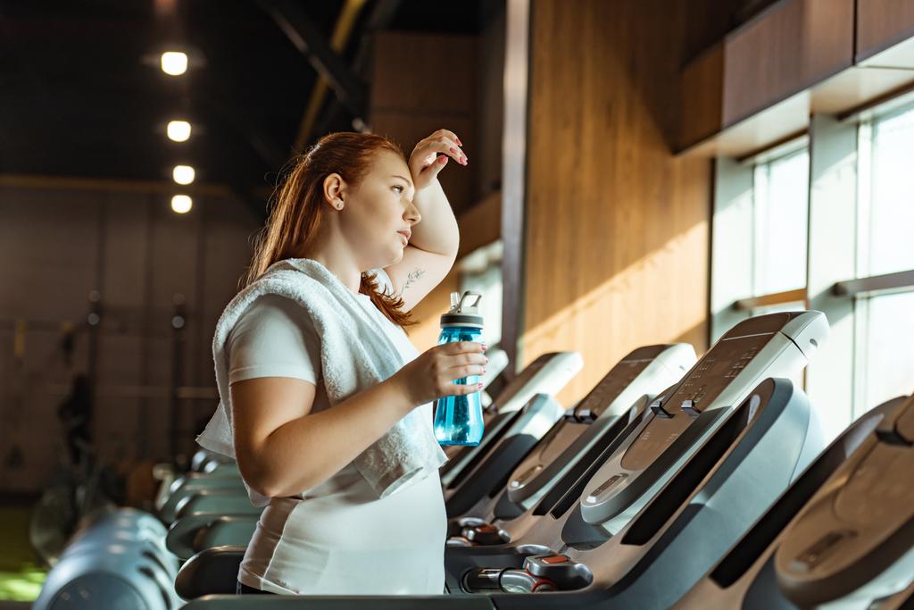 Müdes übergewichtiges Mädchen berührt beim Halten einer Sportflasche auf dem Laufband die Stirn - Foto, Bild
