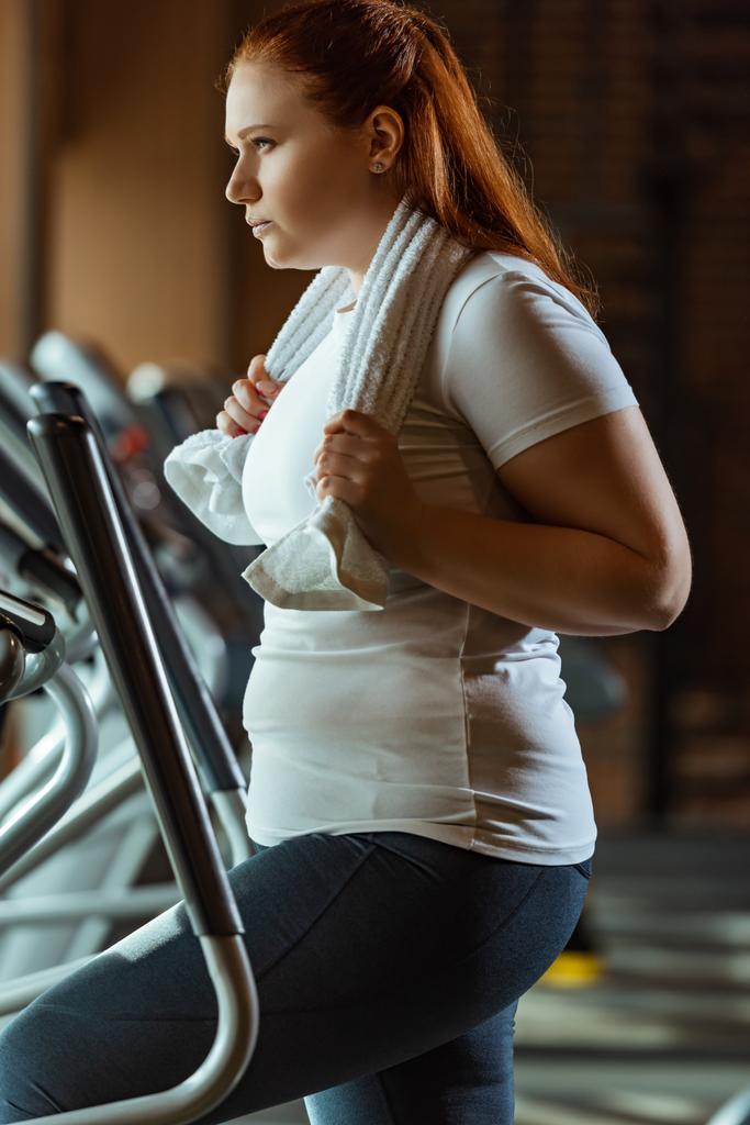 confident overweight girl training on step machine while holding towel - Photo, Image
