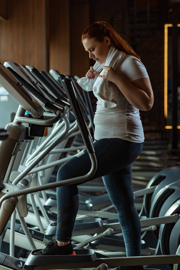 side view of focused overweight girl training on stepper while holding towel - Photo, Image