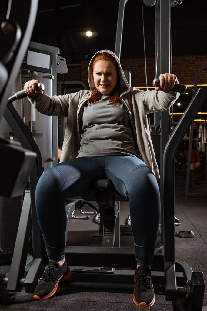 purposeful overweight girl doing arms extension exercise on fitness machine  - Photo, Image