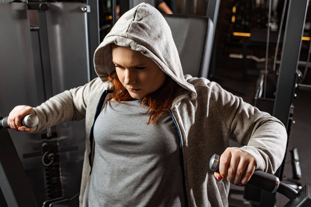 συμπυκνωμένο υπέρβαρο κορίτσι κάνει την επέκταση των όπλων άσκηση στο μηχάνημα γυμναστικής  - Φωτογραφία, εικόνα