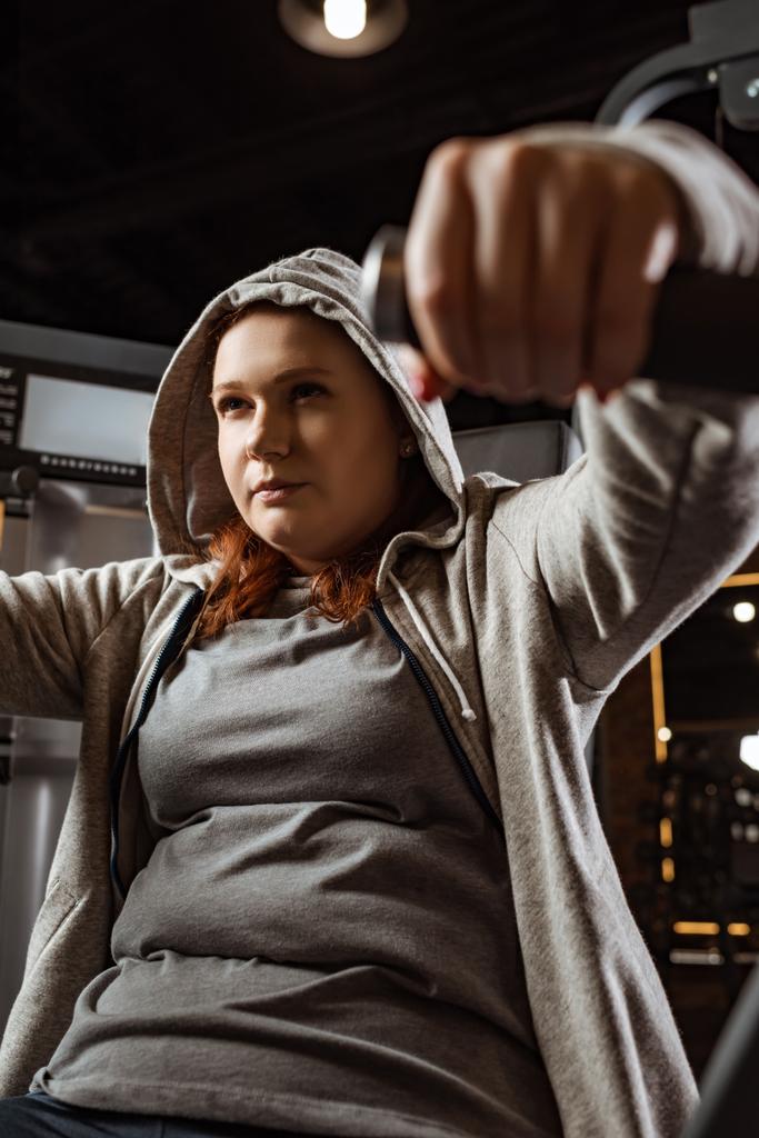 selektiver Fokus eines konzentrierten übergewichtigen Mädchens, das Armstreckübungen an einem Fitnessgerät macht  - Foto, Bild