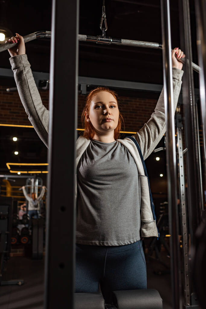 übergewichtiges Mädchen schaut in die Kamera, während sie Armstreckübungen auf einem Fitnessgerät macht - Foto, Bild