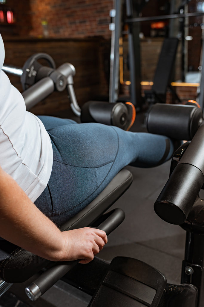 vista recortada de chica con sobrepeso haciendo ejercicio de extensión de pierna en la máquina de fitness
 - Foto, Imagen