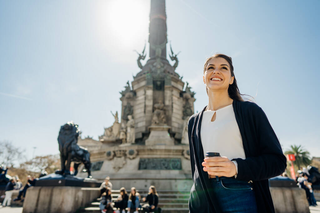 Uśmiechnięta młoda kobieta stojąca przed pomnikiem Kolumba w Barcelonie, Hiszpania.Pijąca kawę w ruchliwe poranki. Turysta podróżujący do Europy.Wakacje w Hiszpanii - Zdjęcie, obraz