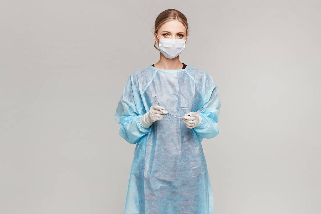 Doctora o cirujana en uniforme médico y máscara protectora médica sosteniendo tubo endotraqueal y posando sobre un fondo gris, aislada. Asistencia sanitaria y emergencia
 - Foto, imagen