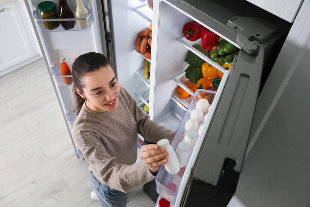 Νεαρή γυναίκα που παίρνει γιαούρτι από το ψυγείο σε εσωτερικούς χώρους, πάνω από την προβολή - Φωτογραφία, εικόνα