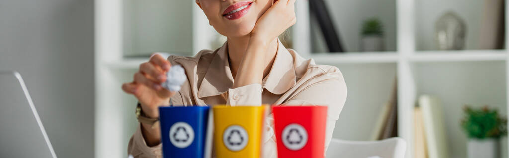 panoramiczne ujęcie uśmiechniętej bizneswoman rzucającej pognieciony papier do wiader na śmieci - Zdjęcie, obraz