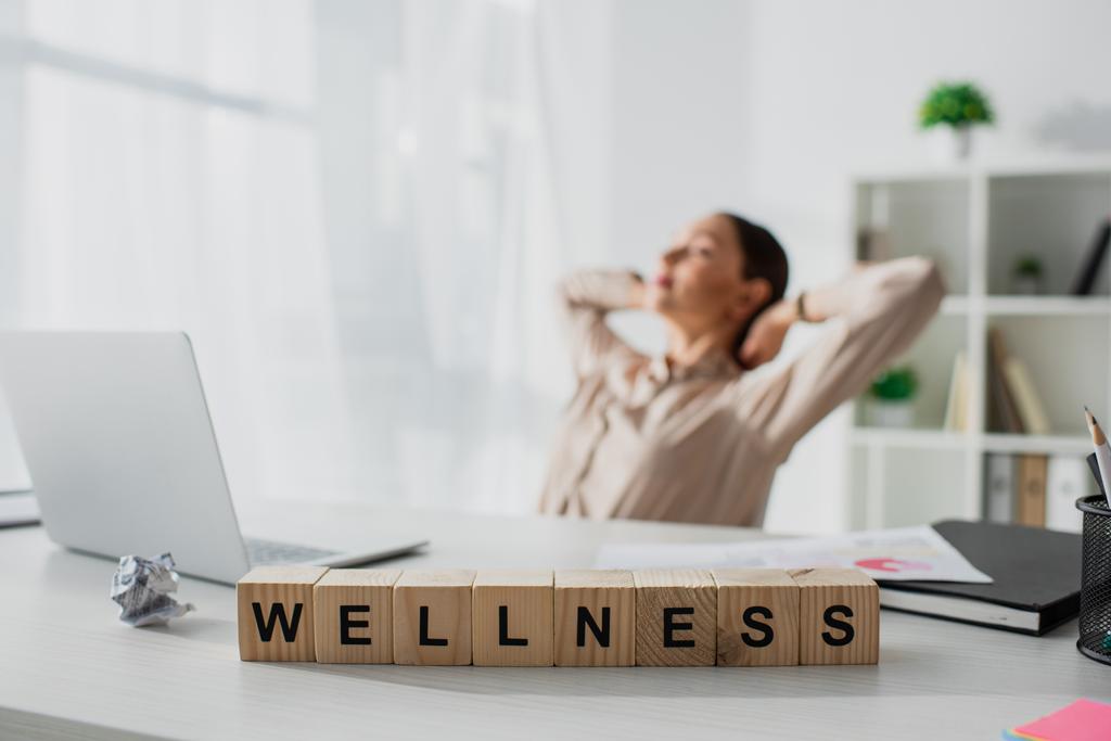 selective focus of businesswoman relaxing at workplace with laptop and alphabet cubes with wellness word  - Photo, Image