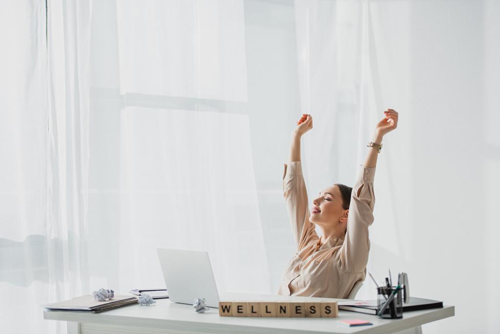 usmívající se podnikatelka odpočívající na pracovišti se zmačkanými papíry, notebookem a abecedními kostkami s wellness slovem  - Fotografie, Obrázek