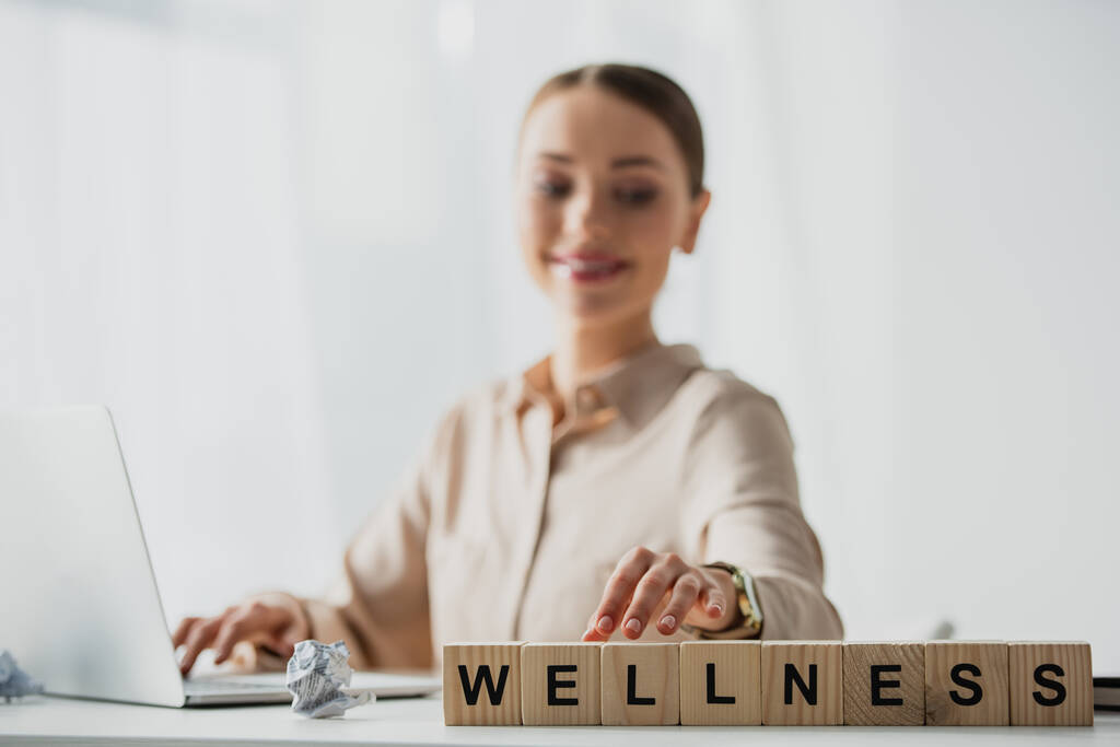selective focus of happy businesswoman working with laptop at workplace with alphabet cubes with wellness word  - Photo, Image
