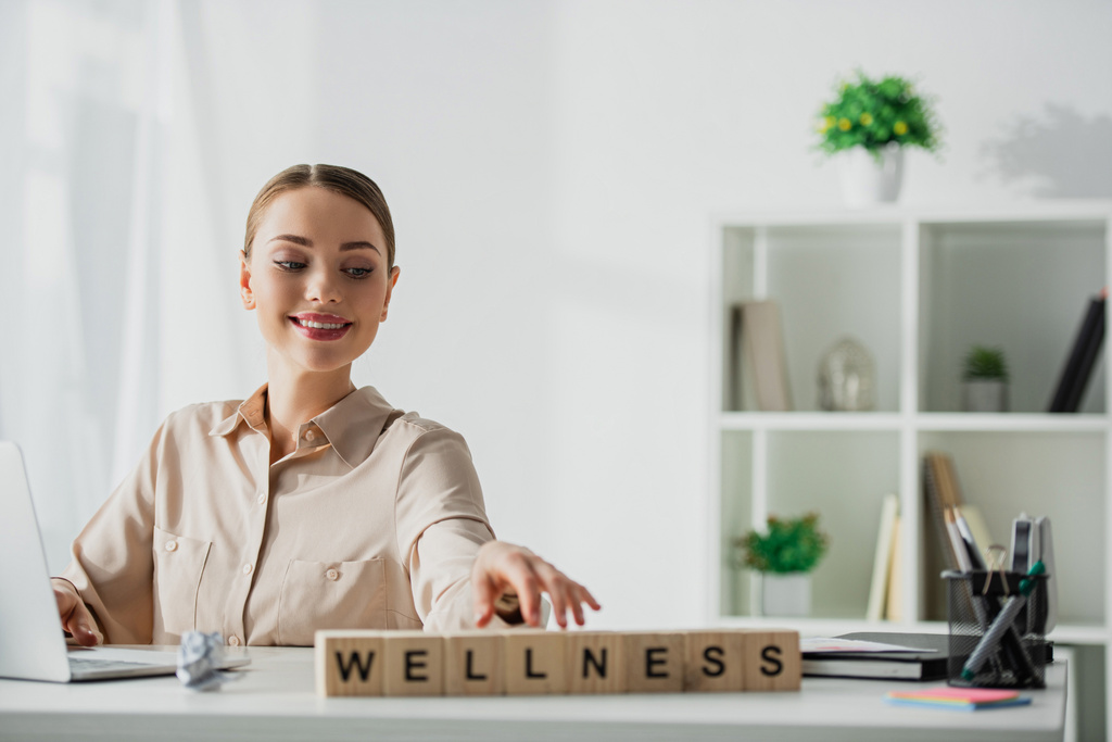 улыбающаяся деловая женщина, сидящая на рабочем месте с ноутбуком и кубиками алфавита со словом wellness
  - Фото, изображение