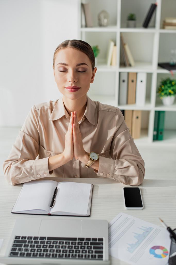 красивая деловая женщина медитирует с namaste жест на рабочем месте с ноутбуком, смартфоном и блокнотом
 - Фото, изображение