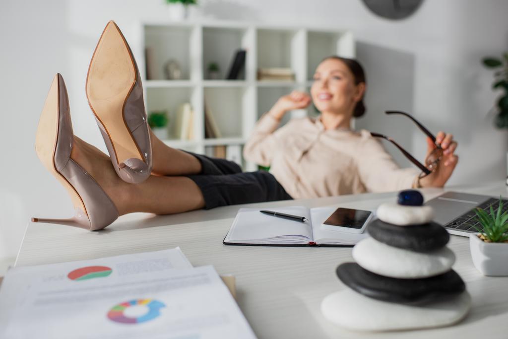 selektivní zaměření podnikatelky sedící s nohama na stole s zenovými kameny v kanceláři - Fotografie, Obrázek