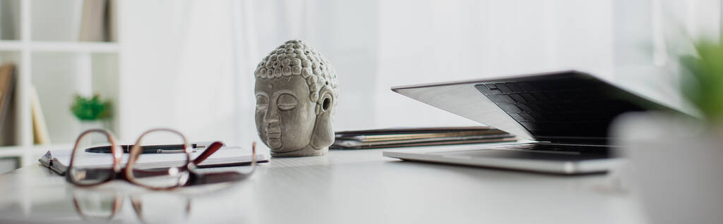 panoramische opname van Boeddha hoofd, bril en laptop op tafel in modern kantoor - Foto, afbeelding