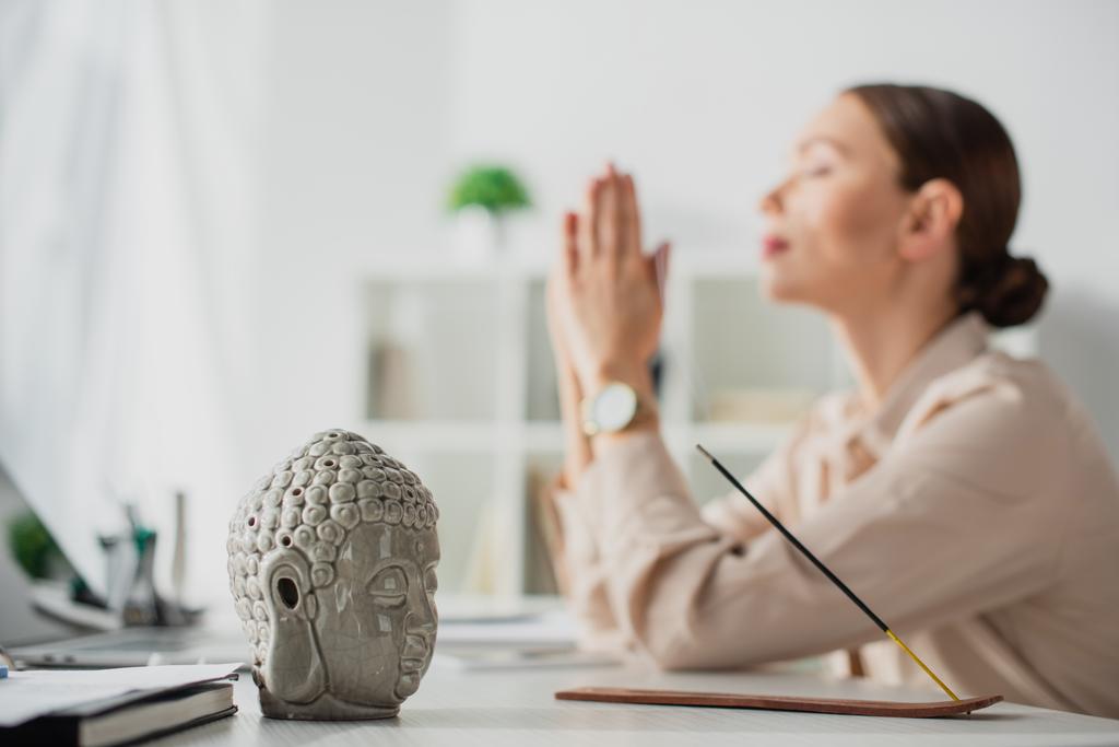 selective focus of businesswoman meditating with namaste gesture at workplace with Buddha head and incense stick - Photo, Image