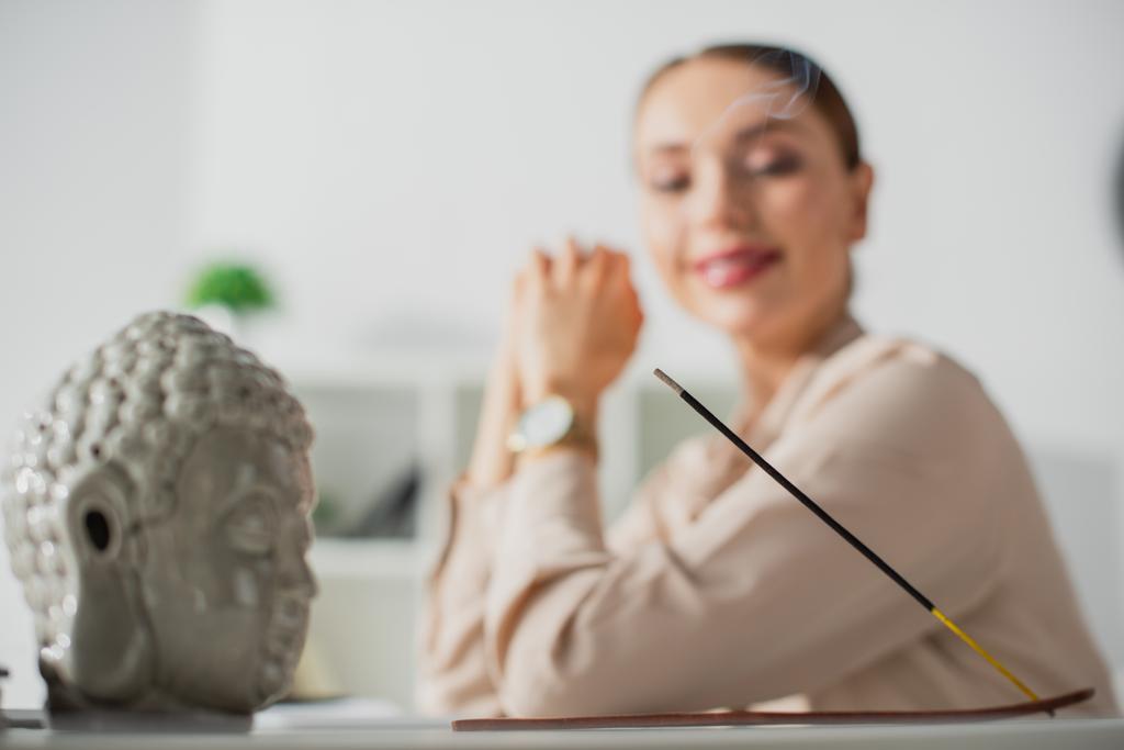 selektivní zaměření šťastné podnikatelky sedící v kanceláři s Buddhovou hlavou a kadidlem - Fotografie, Obrázek