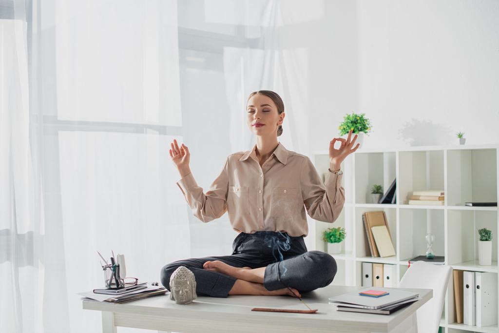 attractive businesswoman meditating in lotus pose with gyan mudra at workplace with Buddha head and incense stick - Photo, Image