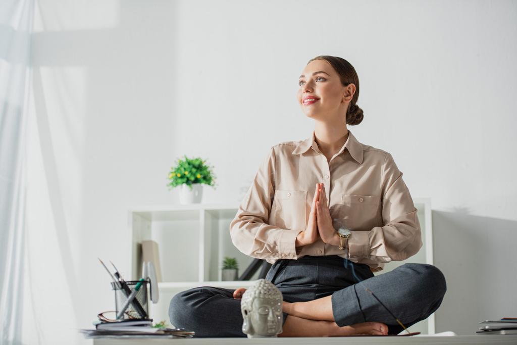 mulher de negócios feliz meditando em pose de lótus com gesto namaste no local de trabalho com a cabeça de Buda e pau de incenso
 - Foto, Imagem