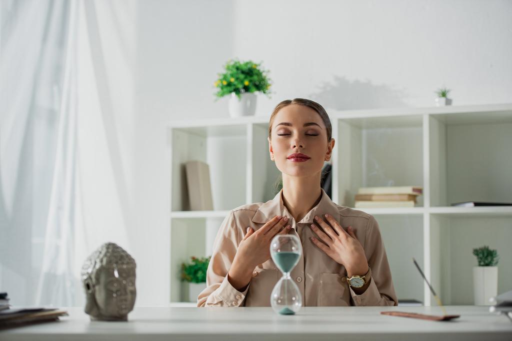 приваблива втомлена бізнес-леді, що сидить на робочому місці з піщаним годинником і головою Будди
 - Фото, зображення