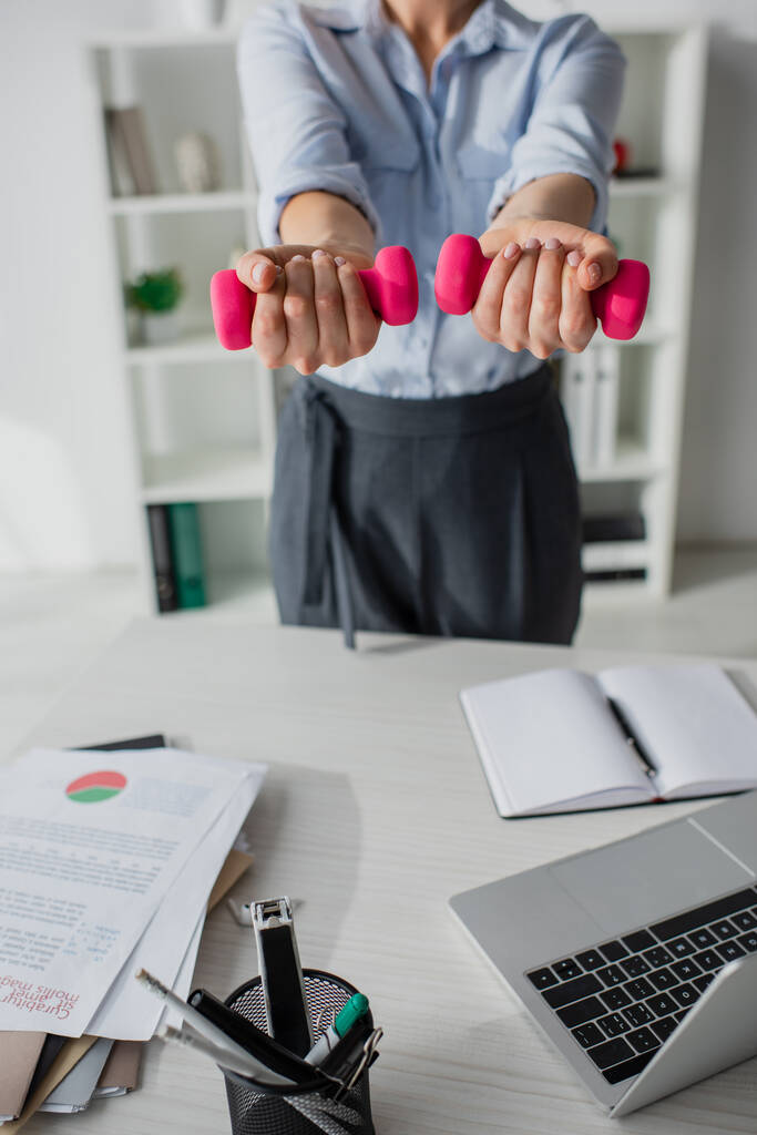 cropped view of businesswoman training with dumbbells at workplace with laptop and paperwork - Photo, Image