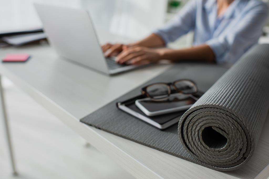 selective focus of businesswoman working on laptop at workplace with fitness mat, notepad, smartphone and eyeglasses - Photo, Image