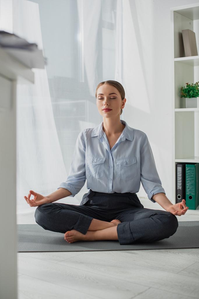 atractiva empresaria practicando yoga en posición de loto con gyan mudra sobre estera en oficina
  - Foto, Imagen