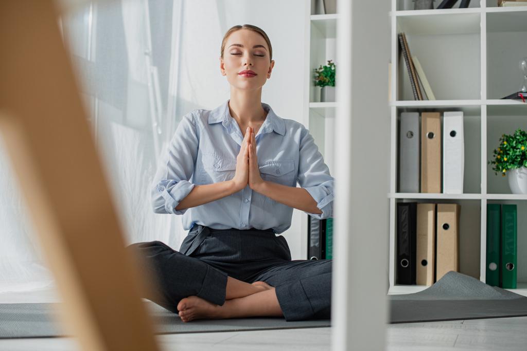 selective focus of businesswoman practicing yoga in lotus position with namaste gesture on mat in office  - Photo, Image