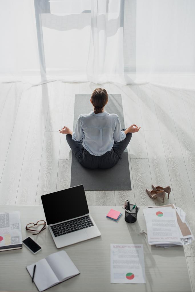 visão traseira de empresária praticando ioga na posição de lótus com mudra gyan no tapete no escritório
  - Foto, Imagem