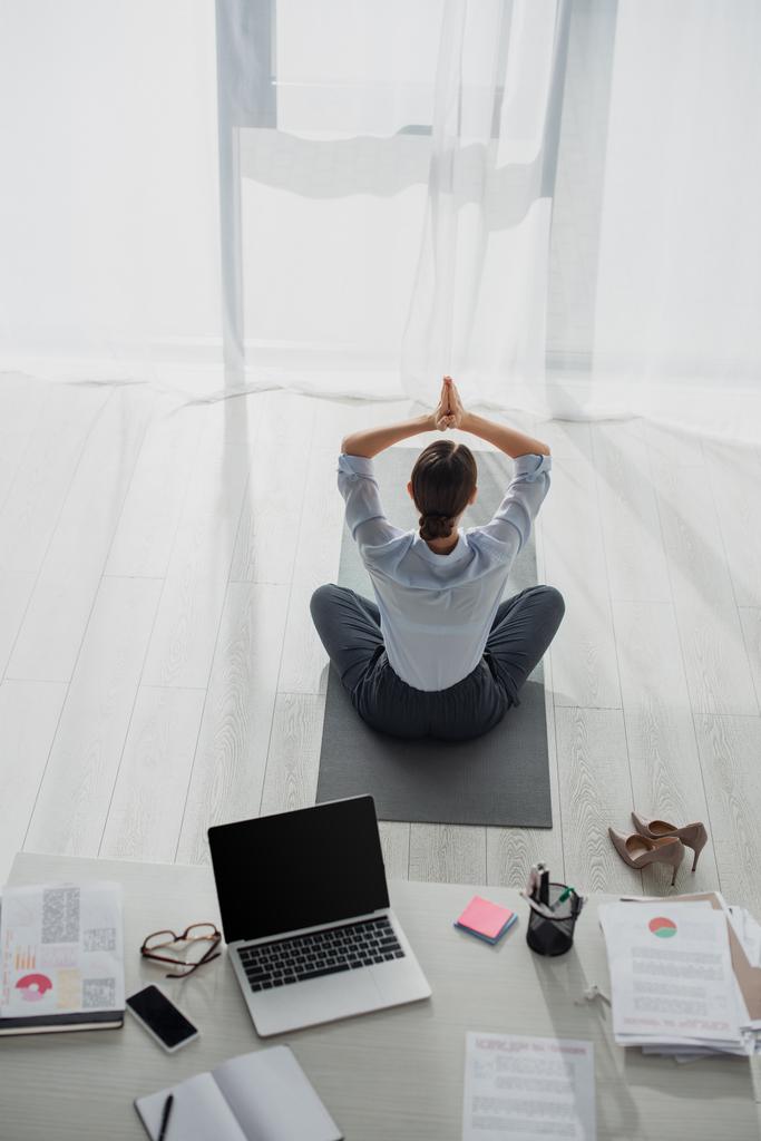vista posterior de mujer de negocios practicando yoga en posición de loto con gesto namaste en estera en oficina con laptop
 - Foto, imagen