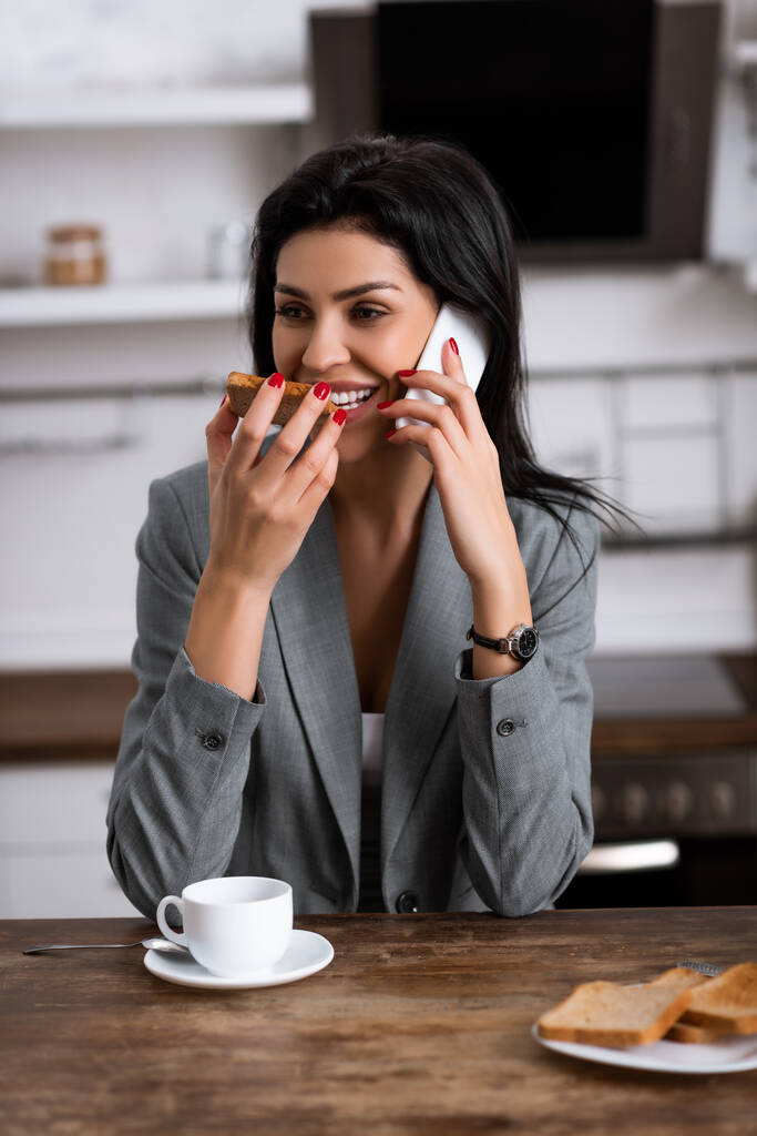 glimlachende zakenvrouw het eten van toast brood met jam tijdens het praten op de smartphone en het verbergen van probleem van huiselijk geweld  - Foto, afbeelding
