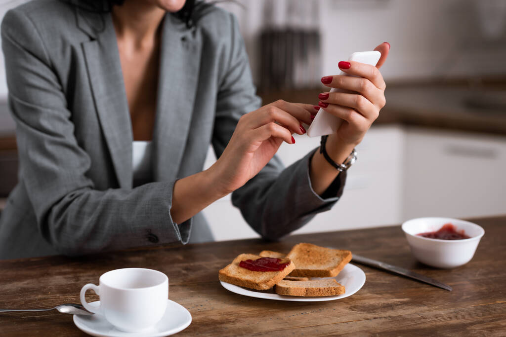 vista ritagliata di donna d'affari utilizzando smartphone vicino a colazione a casa
 - Foto, immagini