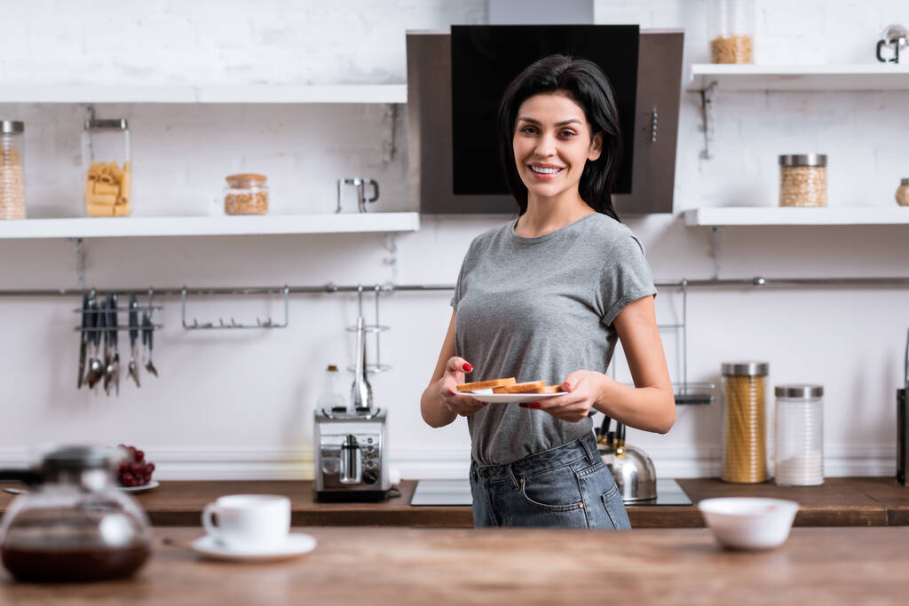 selectieve focus van gelukkige vrouw met bord met toast brood en staan in de keuken  - Foto, afbeelding