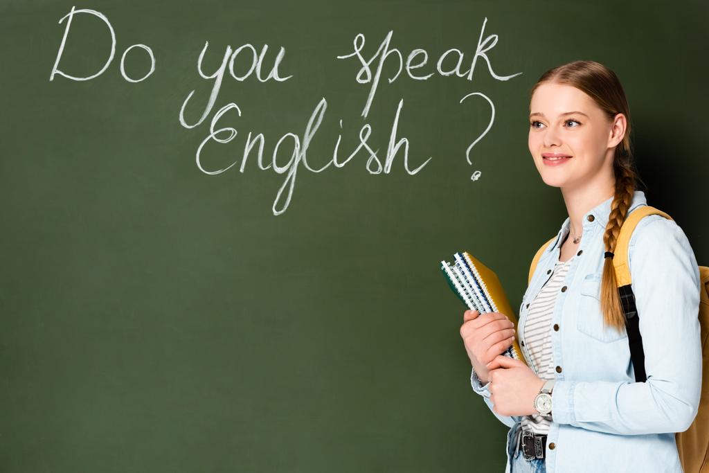usmívající se dívka s copybooks a batohem stojící v blízkosti tabuli s mluvíte anglicky písmo - Fotografie, Obrázek
