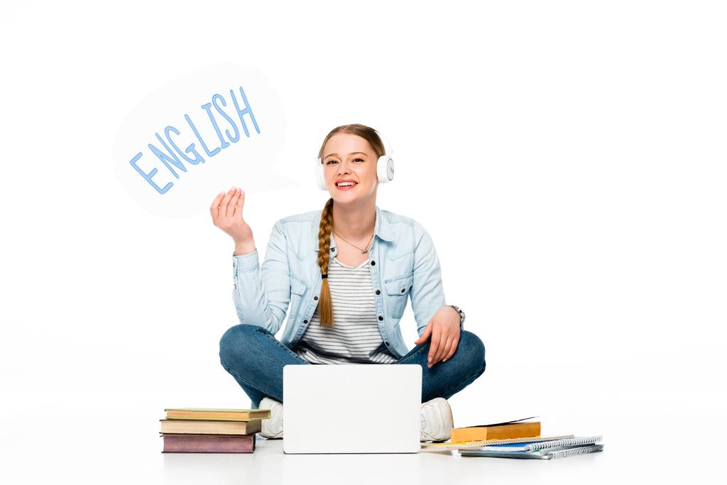 uśmiechnięta dziewczyna siedząca na podłodze w słuchawkach z bańką mowy z angielskim napisem w pobliżu laptopa, książek i copybooków odizolowanych na białym - Zdjęcie, obraz