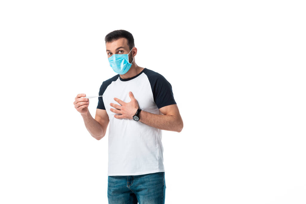 Kranker Mann in medizinischer Maske, digitales Thermometer in der Hand und isoliert auf weißem Grund stehend  - Foto, Bild