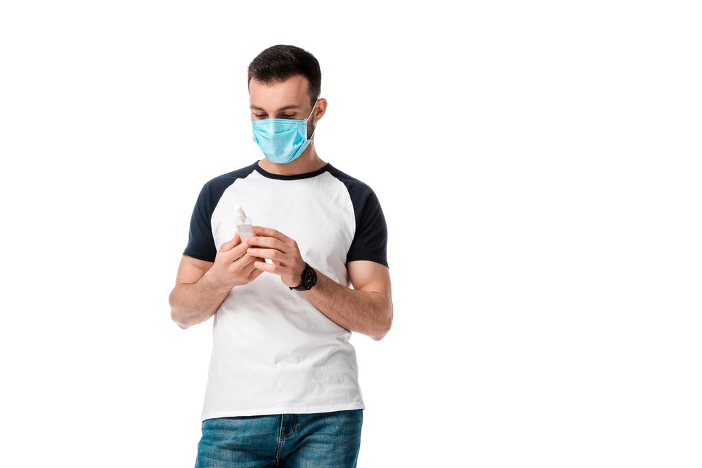 άνθρωπος με ιατρική μάσκα κοιτάζοντας φιάλη ψεκασμού με αντιβακτηριδιακό υγρό που απομονώνεται σε λευκό  - Φωτογραφία, εικόνα