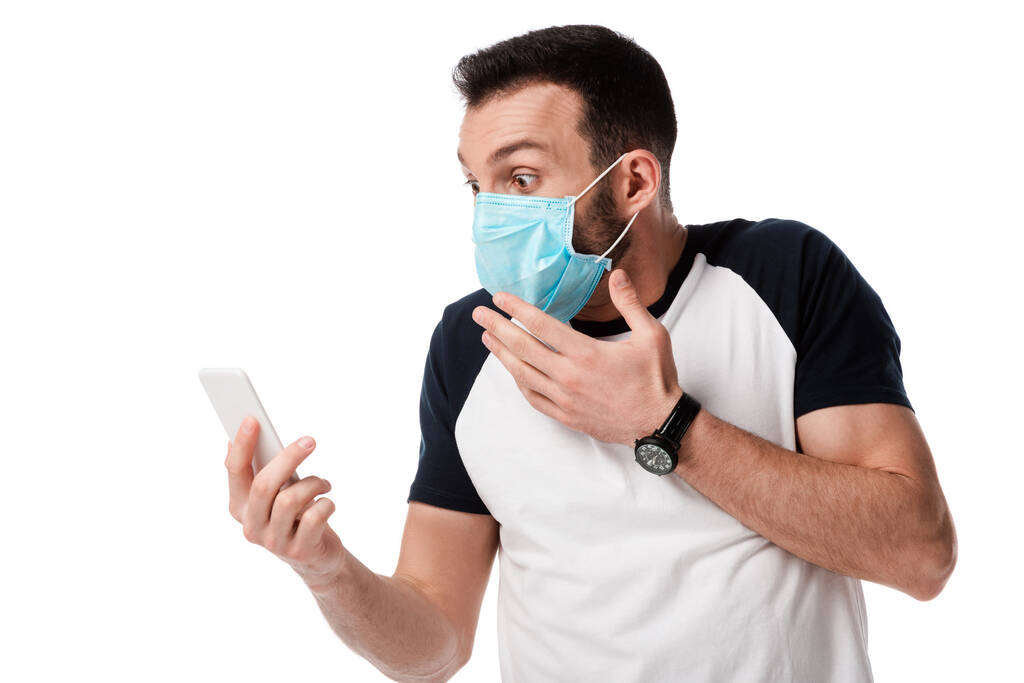 шокований чоловік у медичній масці дивиться на смартфон ізольовано на білому
  - Фото, зображення