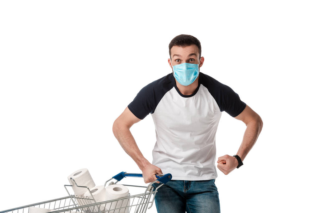 Tıbbi maskeli bir adam, alışveriş arabasının yanında beyaz tuvalet kağıdı olan kasları gösteriyor. - Fotoğraf, Görsel