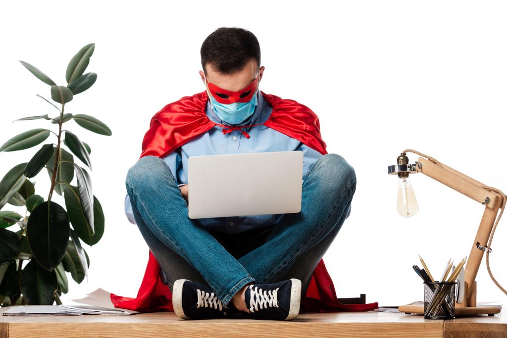 hombre en máscara médica y capa de superhéroe usando el ordenador portátil y sentado en la mesa aislado en blanco
  - Foto, Imagen