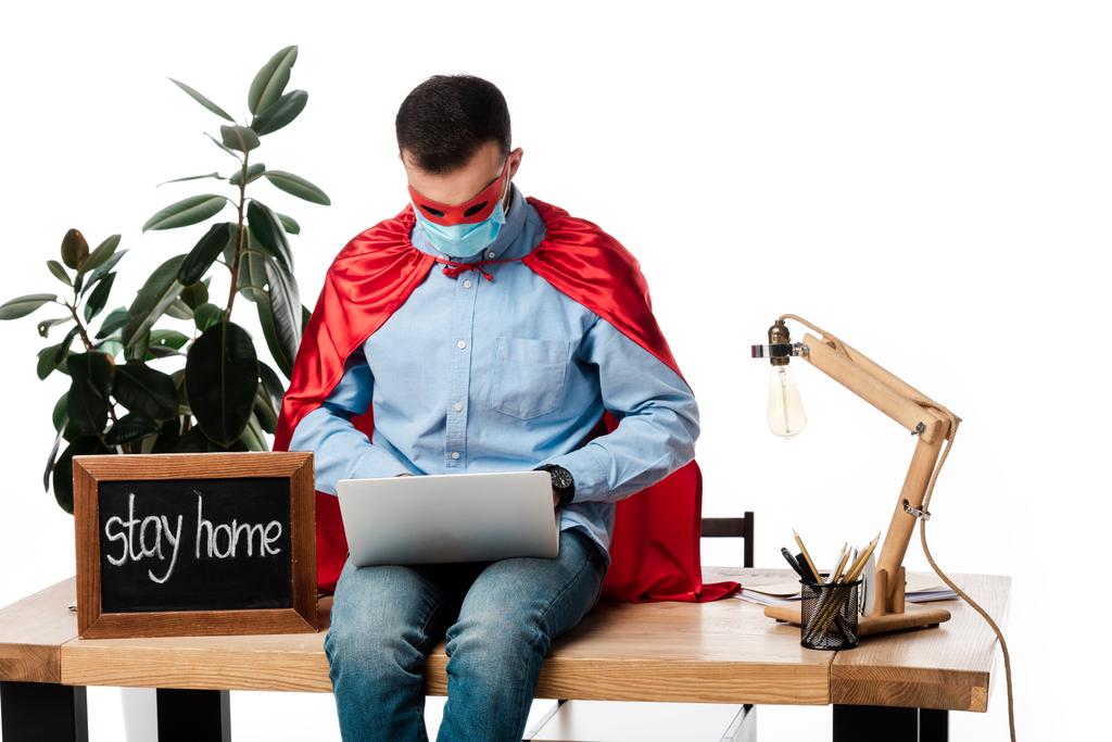 freelancer in medical mask and superhero costume using laptop near chalk board with stay home lettering isolated on white  - Photo, Image