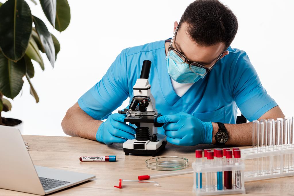 Tıbbi maskeli ve gözlüklü bilim adamı mikroskoba dokunuyor Coronavirus yazıları ve beyaz dizüstü bilgisayarı olan test tüpleri yakınında.  - Fotoğraf, Görsel