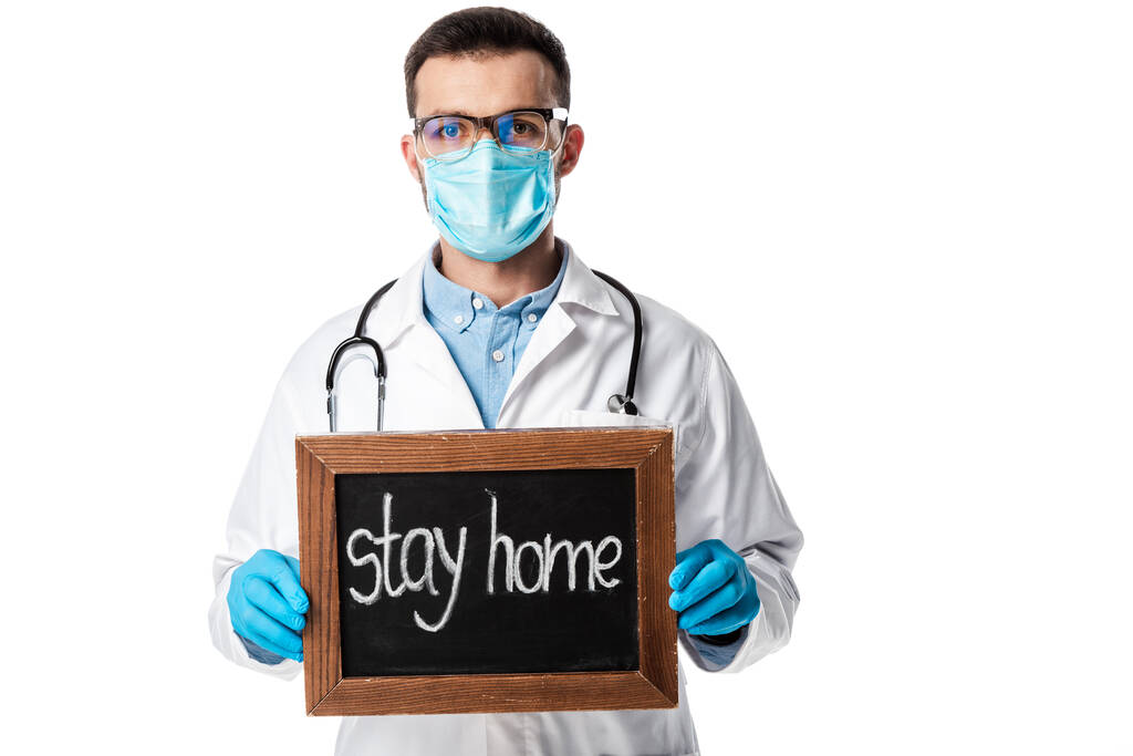 arts in medisch masker, bril en witte jas met krijtbord met verblijf thuis belettering geïsoleerd op wit  - Foto, afbeelding