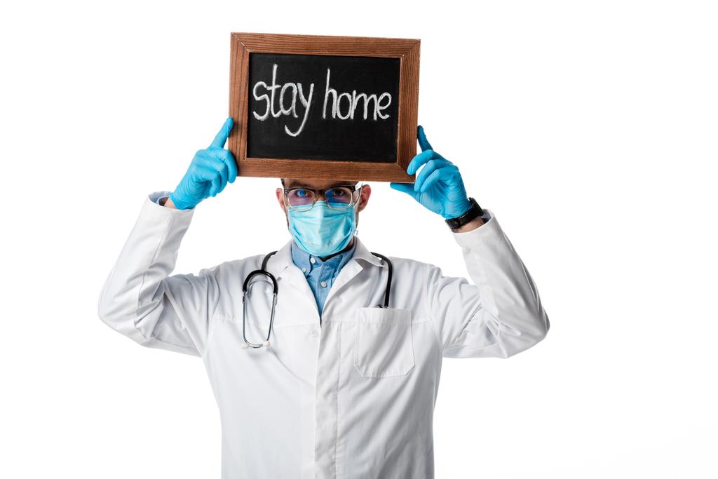 arts in medisch masker en witte jas houden krijtbord met verblijf thuis belettering boven hoofd geïsoleerd op wit  - Foto, afbeelding