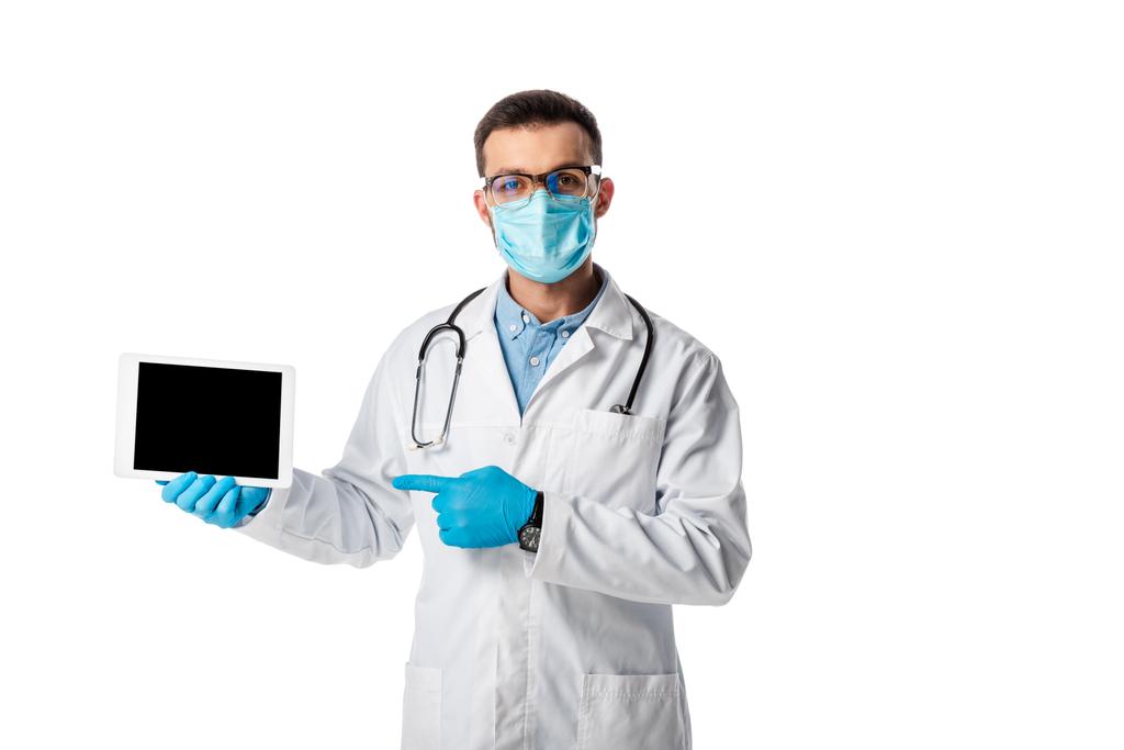 Arzt in medizinischer Maske und weißem Mantel zeigt mit dem Finger auf digitales Tablet mit leerem Bildschirm isoliert auf weißem Hintergrund  - Foto, Bild