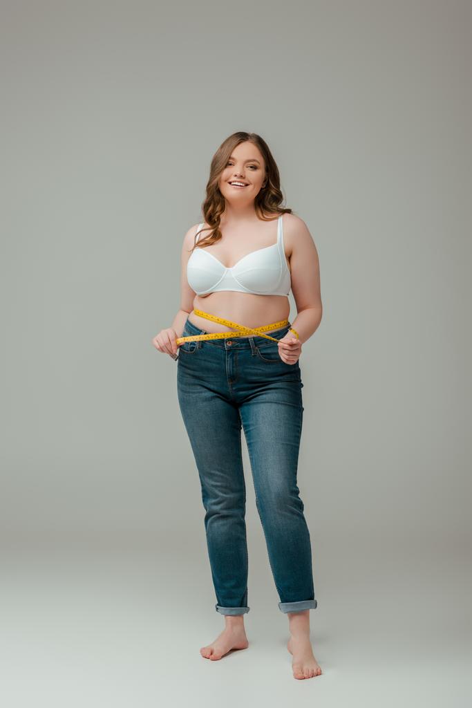 happy plus size woman in jeans and bra measuring waist on grey - Photo, Image