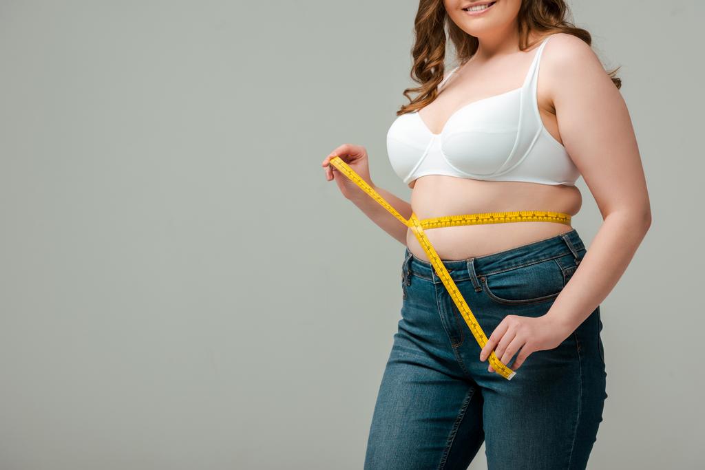vista ritagliata di felice plus size donna in jeans misurazione vita isolato su grigio
 - Foto, immagini