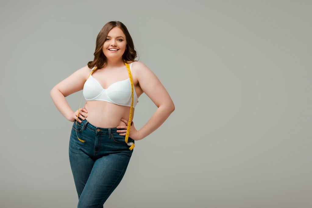 happy plus size woman in jeans and bra standing with hands on hips isolated on grey - Photo, Image